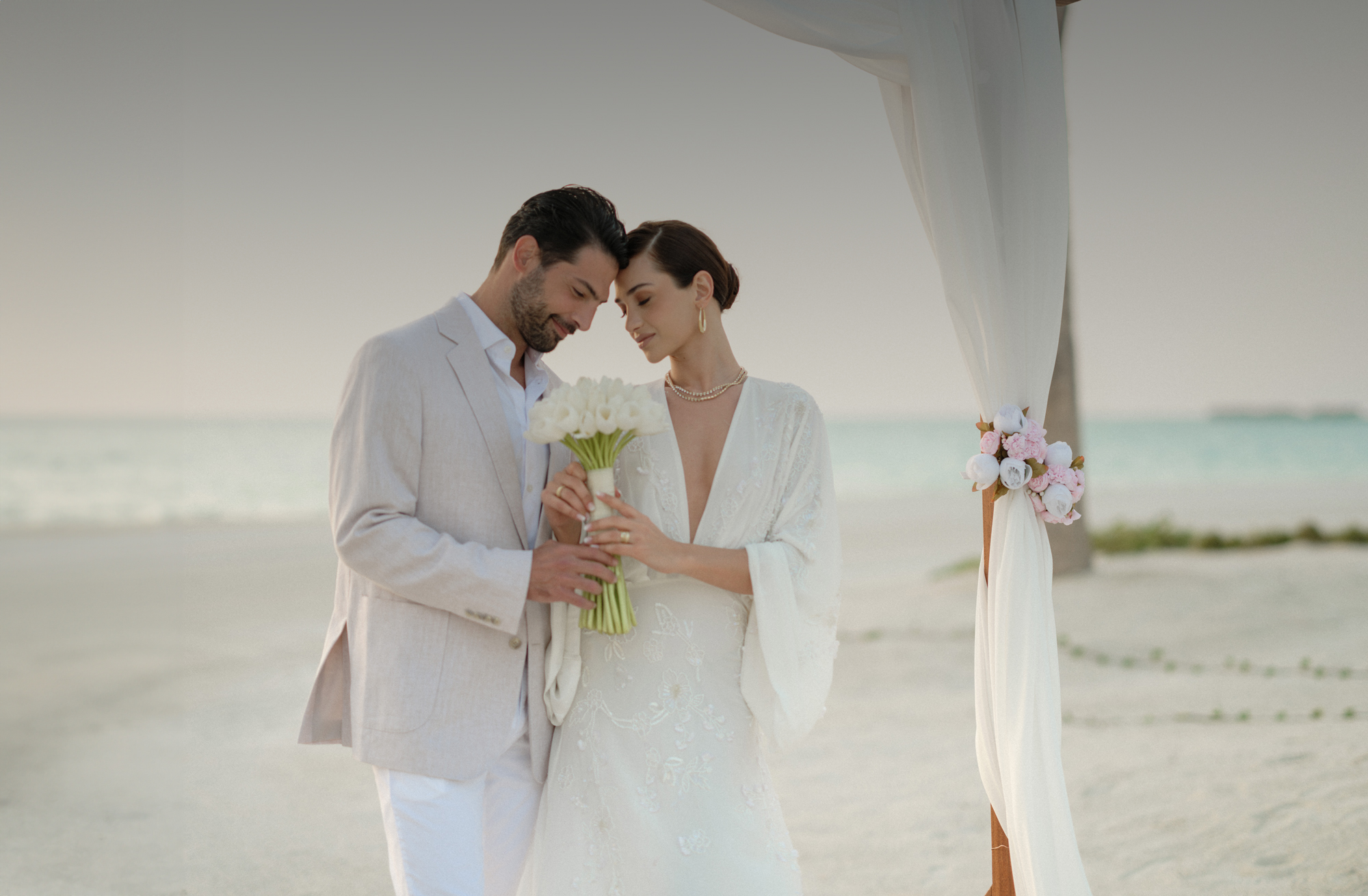 A bride and groom wearing white clothes, with a sunset and beach scene in the background.